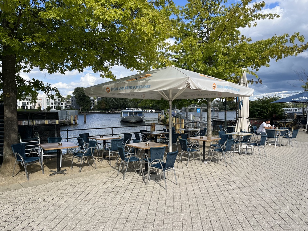 Tisch mit Stühlen unter großem Sonnenschirm direkt am Wasser im Hafen Treptow